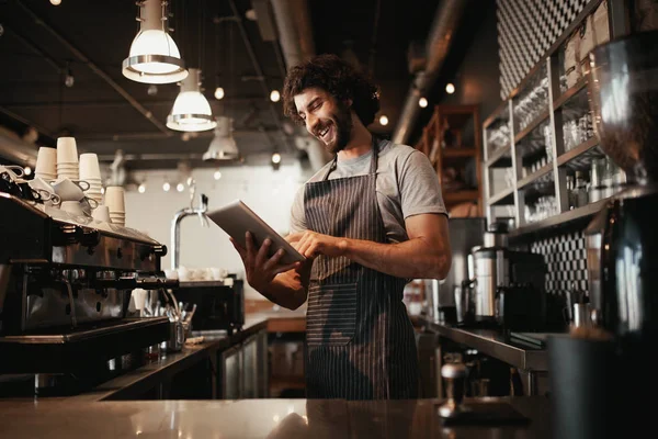 Vrolijke jonge Kaukasische cafe eigenaar draagt zwart gestreepte schort met behulp van digitale tablet — Stockfoto