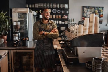 Düşünceli afro-amerikan küçük kahve dükkanı sahibi tezgahın arkasında kollarını kavuşturmuş önlük giymiş başka tarafa bakıyor.
