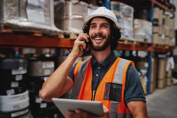 Magazijnmedewerker die digitale tablet gebruikt tijdens een gesprek via mobiele telefoon in een magazijn met een witte helm en een veiligheidsvest — Stockfoto