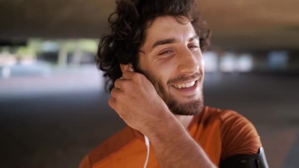 Egy mosolygó sportoló fiatalember portréja, aki zenét hallgat a fehér fülhallgatóján a szabadban. — Stock videók