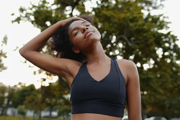 Retrato de uma jovem afro-americana ativa vestindo sutiã esportivo preto com os olhos fechados esticando o pescoço no parque em um dia ensolarado — Fotografia de Stock