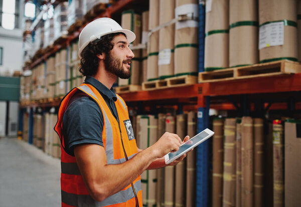 Young warehouse worker in hardhat and safety jacket using digital tablet while taking order and confirming stock availability using digital tablet
