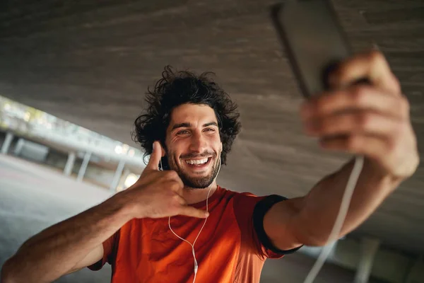 Porträt eines glücklichen jungen Mannes mit Kopfhörer, der mir eine Geste auf seinem Smartphone zuruft — Stockfoto