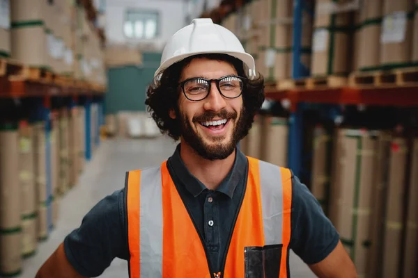 Vrolijke mannelijke opzichter in magazijn met witte helm met oranje vest beschermingspak tussen de planken lachend in een fabriek — Stockfoto