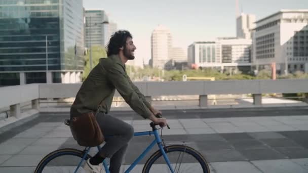 Joven profesional alegre con su bolsa de la frente va a trabajar en bicicleta en la calle de la ciudad — Vídeo de stock
