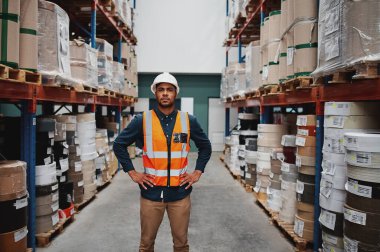 Manager standing in warehouse between shelf filled with goods wearing a white helmet and orange vest for protection with hands on waist