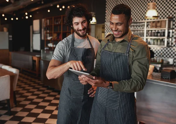 Cheerful cafe partners wearing apron standing inside a coffee shop looking on their touchscreen — ストック写真