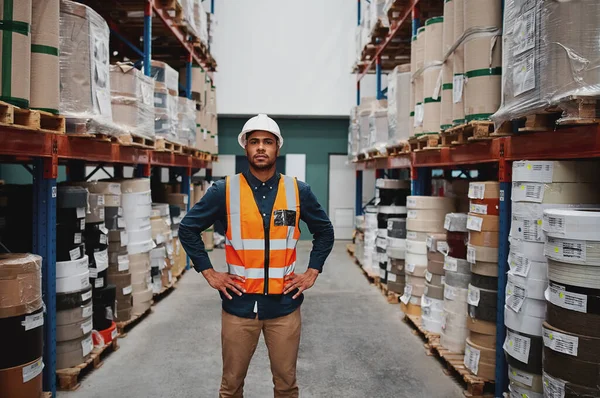 Manager standing in warehouse between shelf filled with goods wearing a white helmet and orange vest for protection with hands on waist — Stockfoto