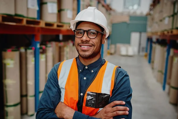 Portrait of handsome cheerful supervisor in warehouse standing with folded hands with material while wearing orange vest and white hardhat