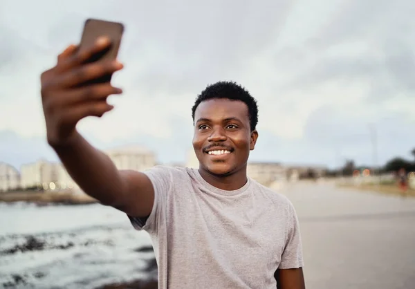 Retrato de um jovem sorrindo em forma de selfie falando em seu telefone inteligente contra o fundo da cidade — Fotografia de Stock