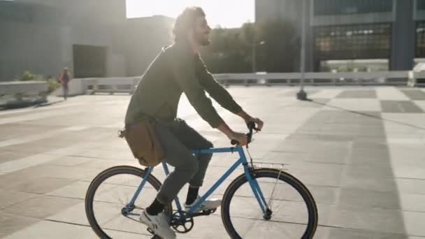 Happy young man with his bag riding bicycle in city street on sunny day — Stockvideo