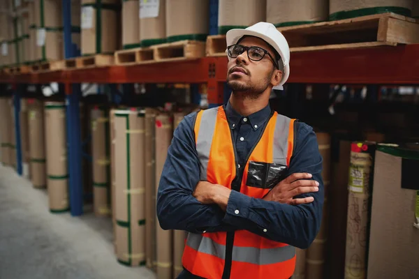 Young warehouse manager standing with arms crossed in factory with white helmet looking away — Stockfoto