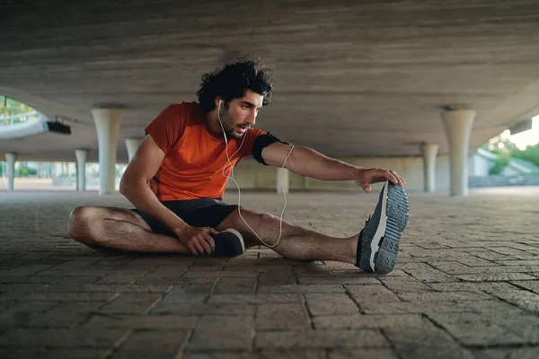 Atleta masculino disfrutando de la música en el auricular calentando y estirando su pierna mientras se relaja bajo el puente de hormigón —  Fotos de Stock