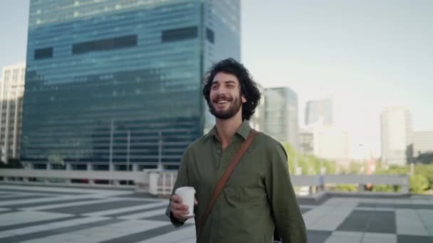 Smiling smart young male professional drinking coffee while walking at downtown with modern corporate building at background — Αρχείο Βίντεο