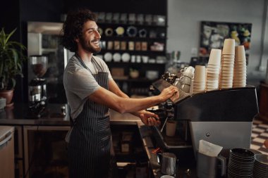 Young cheerful barista wearing black apron while preparing coffee at an automatic machine in a modern coffee shop