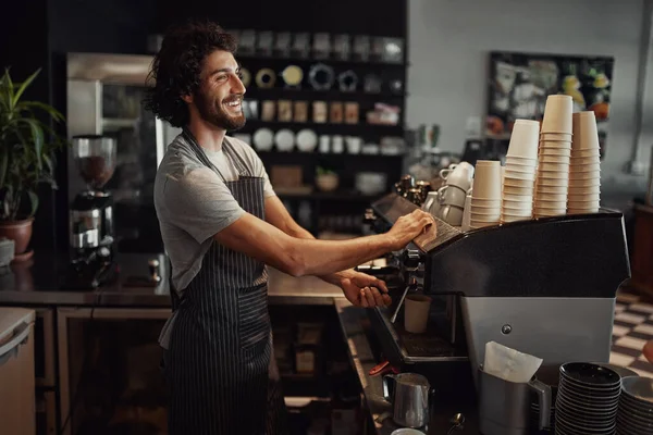 Young cheerful barista wearing black apron while preparing coffee at an automatic machine in a modern coffee shop — Stok fotoğraf