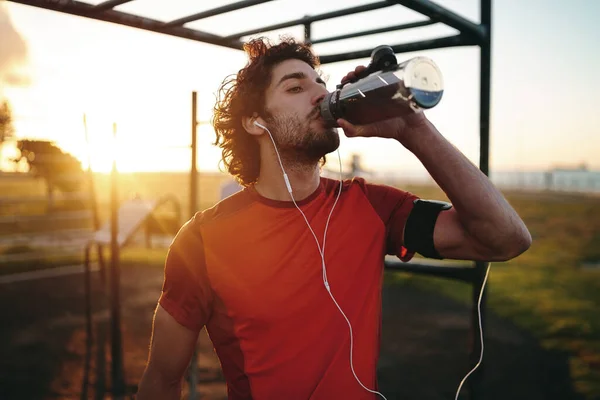 Potret seorang pemuda Kaukasia dengan earphone di telinganya yang sedang minum air setelah berolahraga di taman olahraga terbuka — Stok Foto