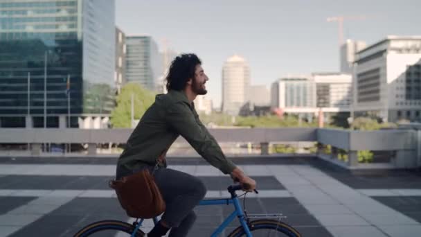 Jovem profissional sorridente bem sucedido andando de bicicleta na frente de edifícios modernos no fundo — Vídeo de Stock