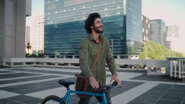 Vista lateral de um jovem empresário sorridente andando ao ar livre perto de edifícios de escritórios com sua bicicleta azul — Vídeo de Stock