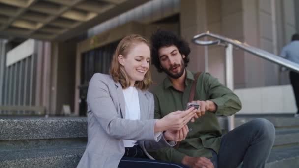 Een aantrekkelijke goed geklede vrouw in zakelijke kleding toont een knappe man haar smartphone samen zitten op kantoor trappenhuis — Stockvideo