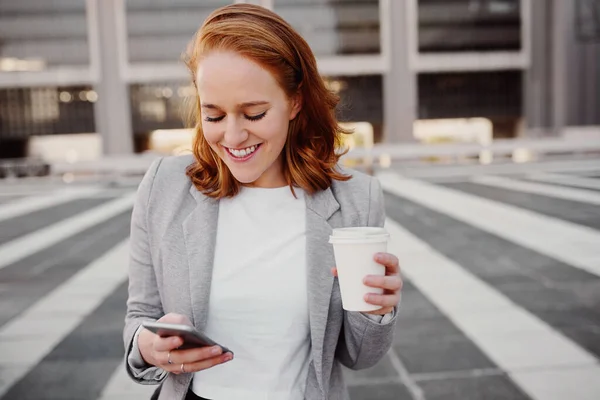Портрет молодої успішної ділової жінки з використанням смартфона, що стоїть на відкритому повітрі, тримає чашку кави — стокове фото