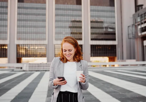 Успішна бізнес-леді за допомогою смартфона, що стоїть на відкритому повітрі, тримає чашку кави — стокове фото