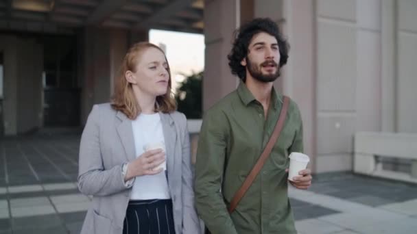 Joven profesional hombre y mujer saliendo del edificio de oficinas sosteniendo una taza de café para llevar sonriendo y hablando entre sí — Vídeos de Stock