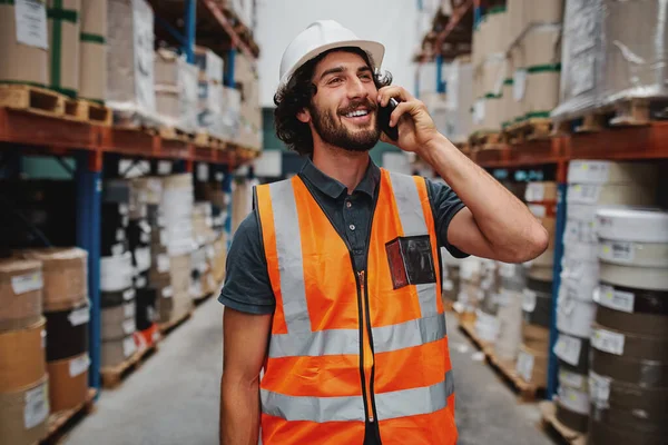 Smiling warehouse manager in conversation on mobile phone wearing white helmet and safety vest standing in between goods shelf looking away — Stock Fotó