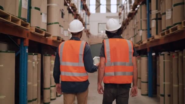 Vue arrière de deux hommes marchant dans l'entrepôt vérifiant les produits manufacturés sur la tablette numérique — Video