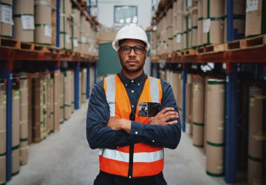 Confident young manager with arms crossed standing wearing orange vest and white hardhat in manufacturing unit looking at camera