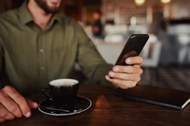 Closeup of male hands using mobile phone drinking coffee while sitting in cafeteria