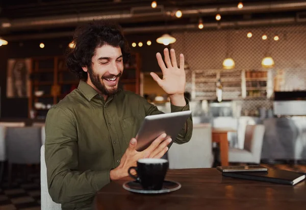 Випадковий бородатий чоловік махає рукою під час розмови через відео-чат у смартфоні, розслабляючись у кафе — стокове фото
