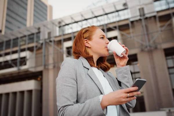 Vista a basso angolo di giovane donna d'affari che tiene smartphone mentre beve caffè dalla tazza monouso bianca — Foto Stock