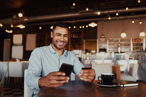 Happy african man holding mobile phone typing card data to make online payment sitting in cafe — Stockfoto
