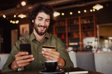 Kafe 'de otururken internet üzerinden ödeme yapmak için cep telefonu daktilo kartı bilgilerini tutan mutlu Kafkas sakallı adam.