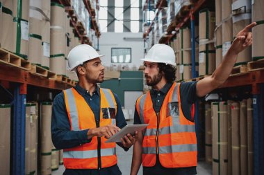 Tensed worker in factory showing shelves to his manager holding digital tablet with hardhat and safety vest