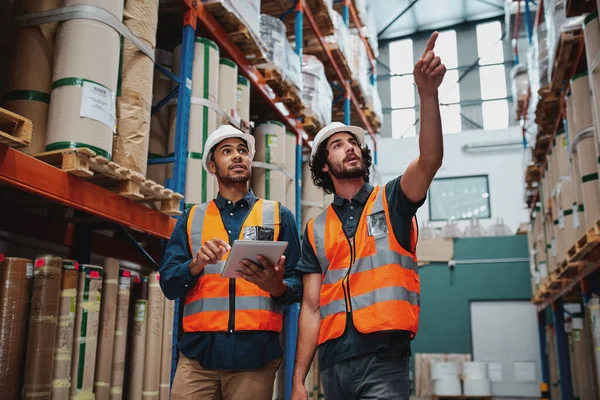 Giovane operaio caucasico che mostra scaffali al suo manager africano in un magazzino con tablet digitale con hardhat e gilet di sicurezza — Foto Stock