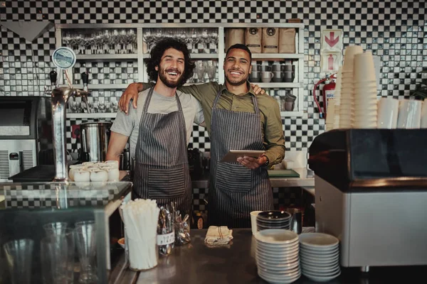 Portrait of smiling colleagues standing behind counter in cafe holding digital tablet with hands on each other shoulder looking at camera — Stock Photo, Image