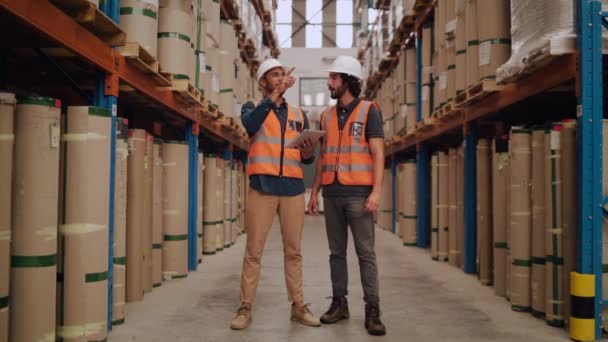Два молодых работника мужского пола, стоящие на складе дистрибуции в белой каске и светоотражающей куртке, осматривая акции с помощью цифрового планшета — стоковое видео