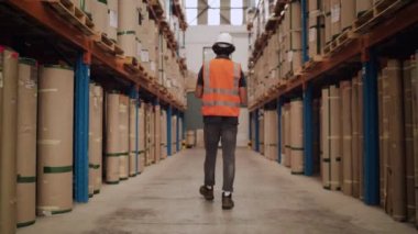 Beyaz kasketli ve yansıtıcı ceketli bir erkek işçinin depodaki büyük paket yığınları arasında yürürken görüntüsü.