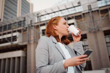 Low-angle view of young businesswoman holding smartphone while drinking coffee from white disposable cup