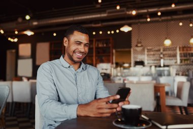 Cheerful handsome african young man relaxing in modern cafe using mobile phone while drinking coffee