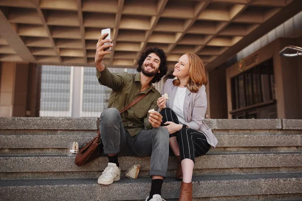 Alegre pareja joven en ropa formal tomando selfie usando teléfono inteligente mientras come sándwich para el almuerzo durante el descanso — Foto de Stock