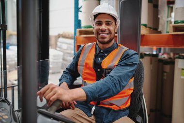 Beyaz miğfer ve yelek giyen mutlu sürücünün portresi forklift makinesinde oturmuş depoda malları taşıyordu.