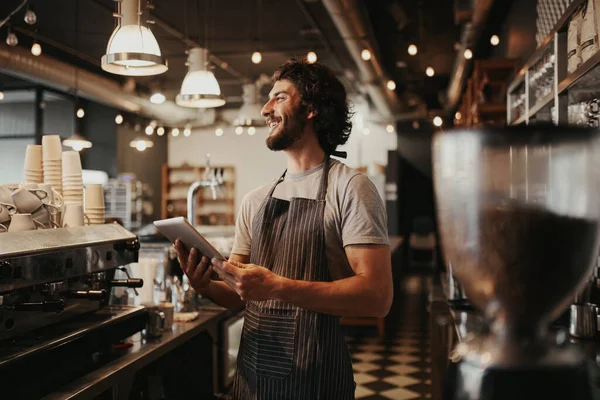 Handsome caucasian man in coffee shop laughing while holding digital tablet in hand standing behind counter