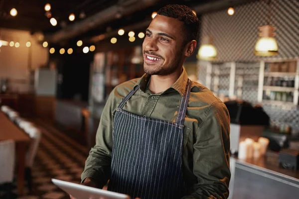 Handsome young man wearing apron holding digital tablet standing in coffee house looking away — Stockfoto