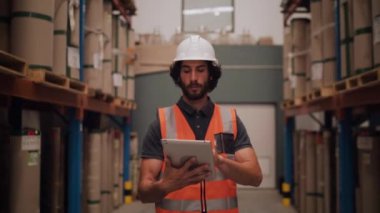 Depodaki erkek işçi, büyük dağıtım malzemelerinin arasında yürüyen dijital tablet listesiyle karşılaştırıyor.
