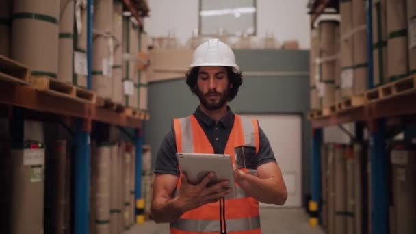 Depodaki erkek işçi, büyük dağıtım malzemelerinin arasında yürüyen dijital tablet listesiyle karşılaştırıyor. — Stok video