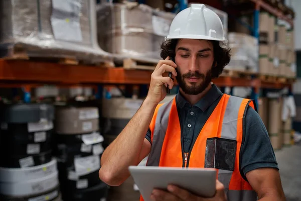 Focused warehouse manager tracciamento dettagli dell'ordine di fornitura utilizzando tablet digitale mentre in conversazione sul telefono cellulare in magazzino — Foto Stock