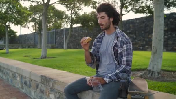 Портрет молодого человека, сидящего со скейтбордом и поедающего сэндвич на обед в парке — стоковое видео